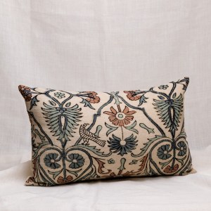 Cojín de color natural con motivos geométricos y flores en tonos azules y tierras, y trasera color natural con dibujo de espiga